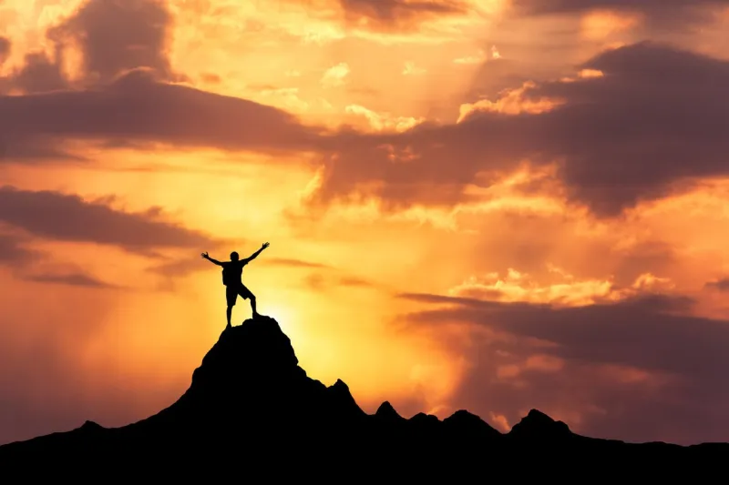 landscape with silhouette of a standing happy man with backpack and raised-up arms on the mountain peak on the background of cloudy sky at colorful sunset in summer travel, climbing, trekking
