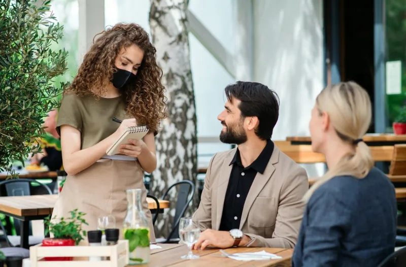 woman waitress with face mask serving happy couple outdoors on terrace restaurant