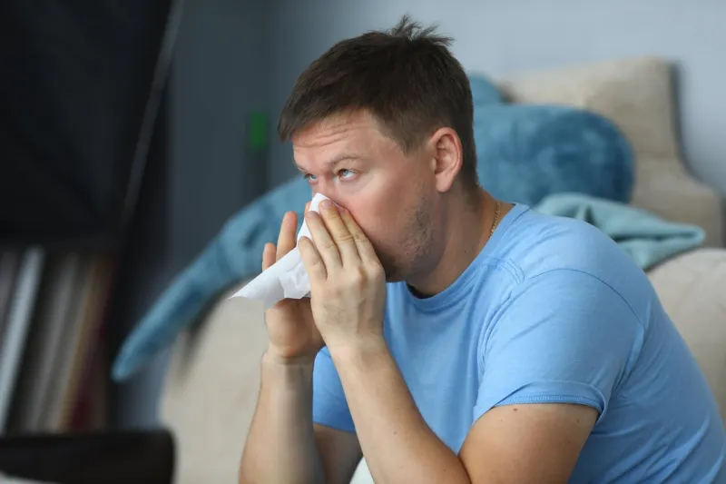 man blowing his nose in a napkin while sitting at home on sofa portrait