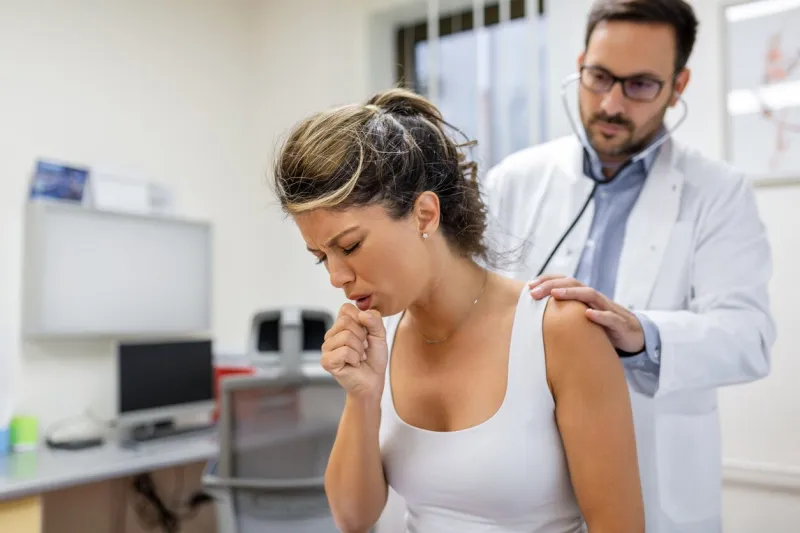 young female patient in the clinic suffered from pneumonia, she is coughing the doctor listens to the wheezing in the lungs with a stethoscope