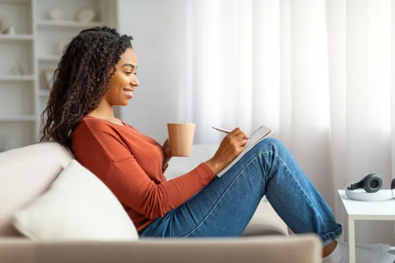 happy young black lady drinking coffee and writing in notepad while relaxing at home, smiling african american woman enjoying cup of tea and taking notes, filling diary or making checklist, side view
