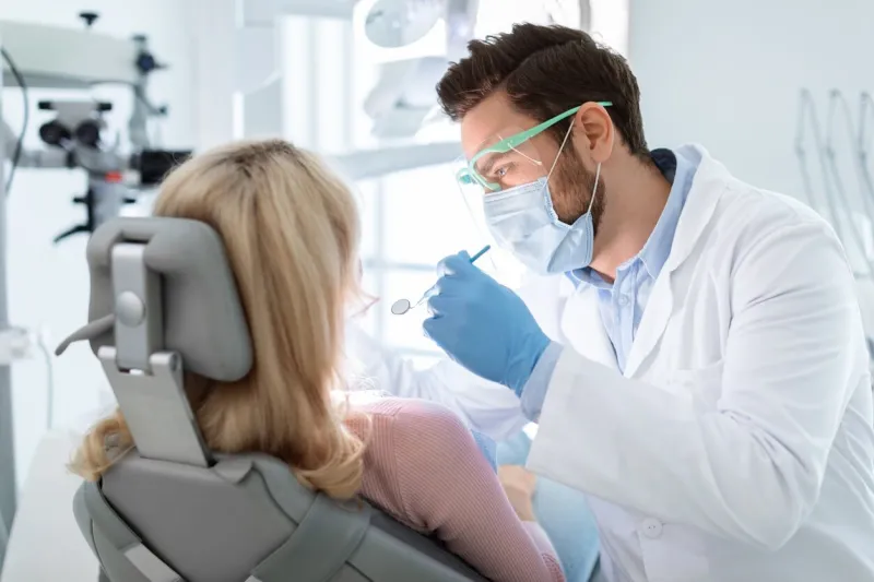 man dentist in face mask and glasses doing treatment for patient blonde lady, holding dental tools, wearing rubber gloves stomatology, dentistry, modern dental clinic concept