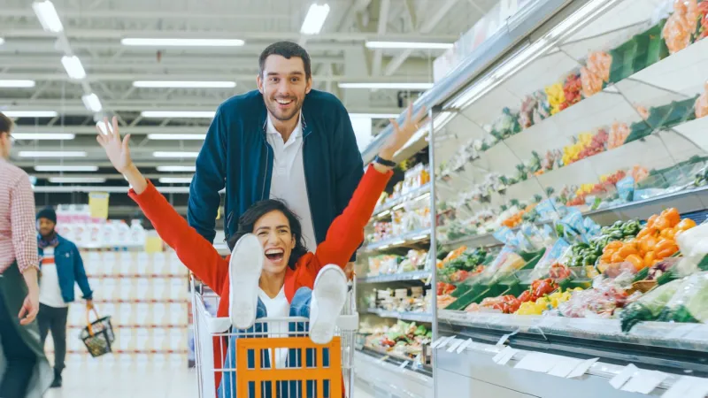 at the supermarket  man pushes shopping cart with woman sitting in it, happy couple has fun racing in a trolley through the fresh produce section of the store people walking by