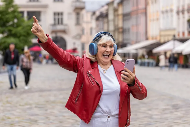 happy overjoyed senior woman in wireless headphones listening favorite energetic music, celebrating win elderly grandmother tourist traveler walking in urban city street town lifestyles outdoors