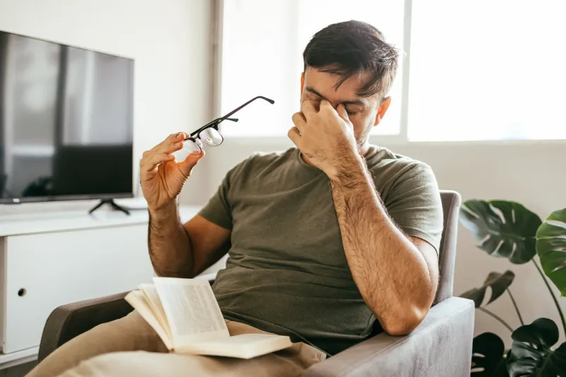 tired man takes off his glasses and rubs his eyes while reading a book at home