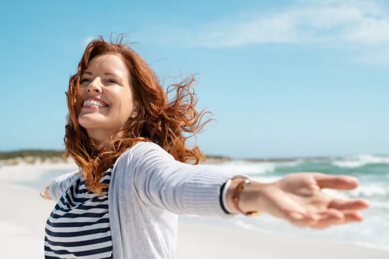 happy mature woman with arms outstretched feeling the breeze at beach beautiful middle aged woman with red hair and arms up dancing on beach in summer during holiday mid lady in casual feeling good and enjoying freedom with open hands at sea, copy space