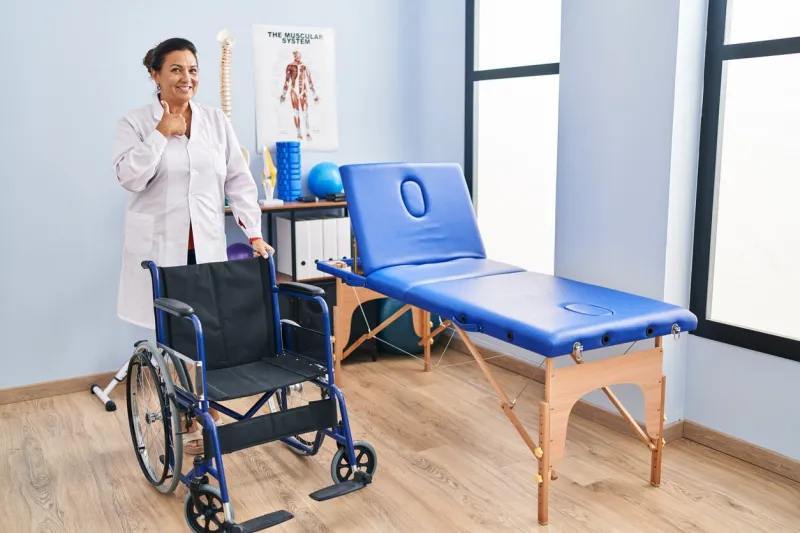 middle age hispanic woman with wheelchair at physiotherapy clinic pointing finger to one self smiling happy and proud