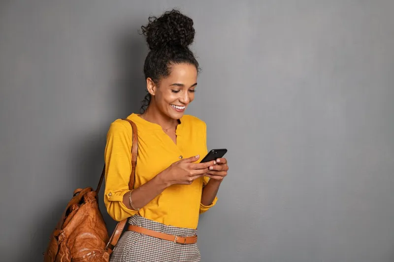 young beautiful african woman with leather backpack standing on grey wall while using smartphone stylish latin girl isolated against gray background checking mobile phone with copy space portrait of smiling tourist using app cellphone