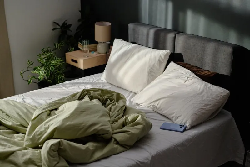 high angle shot of crumpled bed lit with bright sunrays, pastel blue smartphone next to pillow