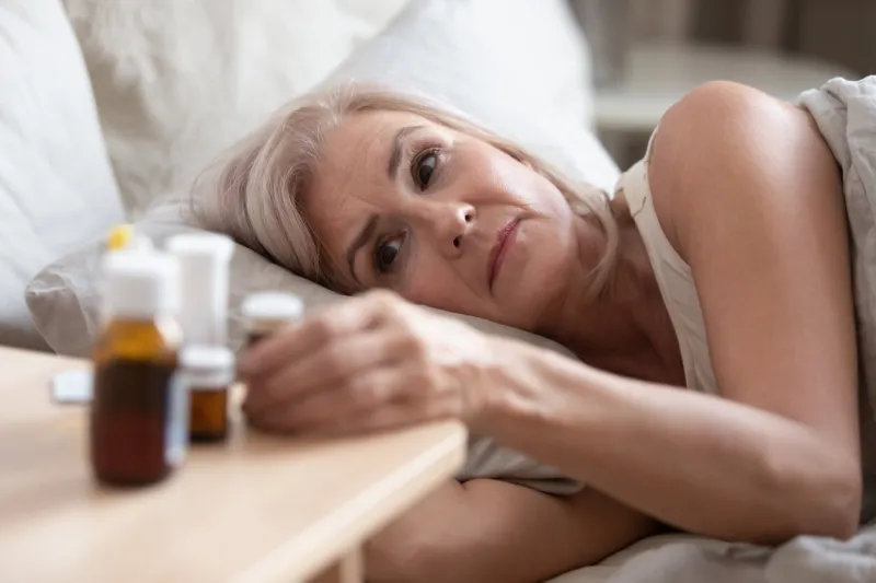close up unhealthy old woman lying in bed woke up in morning looking at heap bottle of pills on bedside table feels cheerless, tablets to reduce menopause symptoms, chronic disease treatments concept