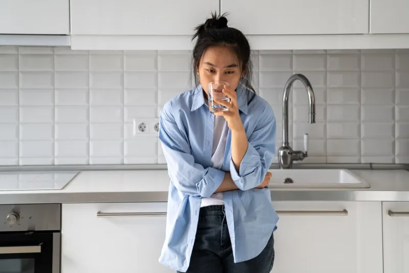 unhappy disheveled asian girl drinks water from glass while standing in kitchen waking up late morning bad mood, drinking an antidepressant korean middle-aged sick woman indigestion and thirsty