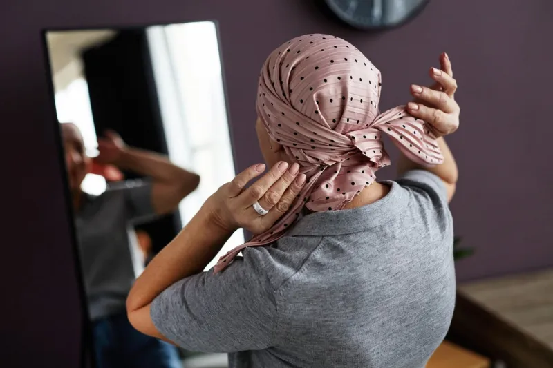 back view of unrecognizable bald woman tying headscarf looking in mirror