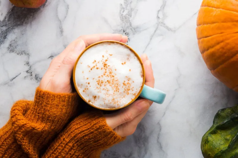 autumn moody background with mug of latte coffee and pumpkins on marble table flat lay in fall colors female hands in cozy sweater