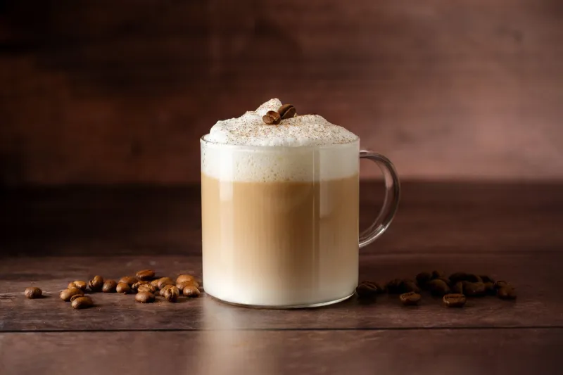 glass cup of layered coffee drink with milk foam, coffee beans over wooden background, copy space