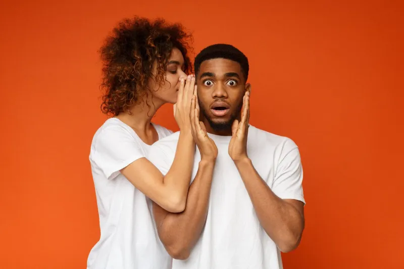 hidden desires black woman whispering her dreams to shocked boyfriend, orange studio background
