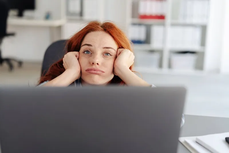 bored young businesswoman pulling a face and pouting as she rests her chin on her hands and peers over her computer at the camera