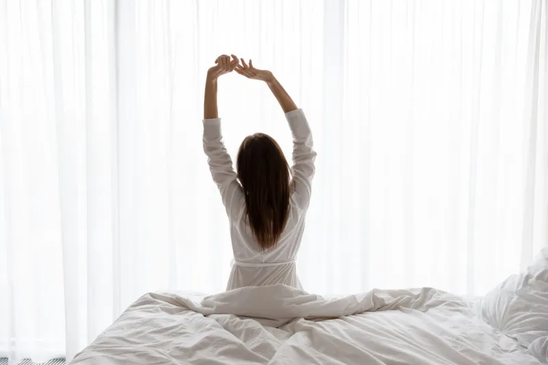 rear view beautiful woman wearing white bathrobe doing exercise, sitting on comfortable bed in modern apartment or hotel, young female stretching arms in early morning after awakening