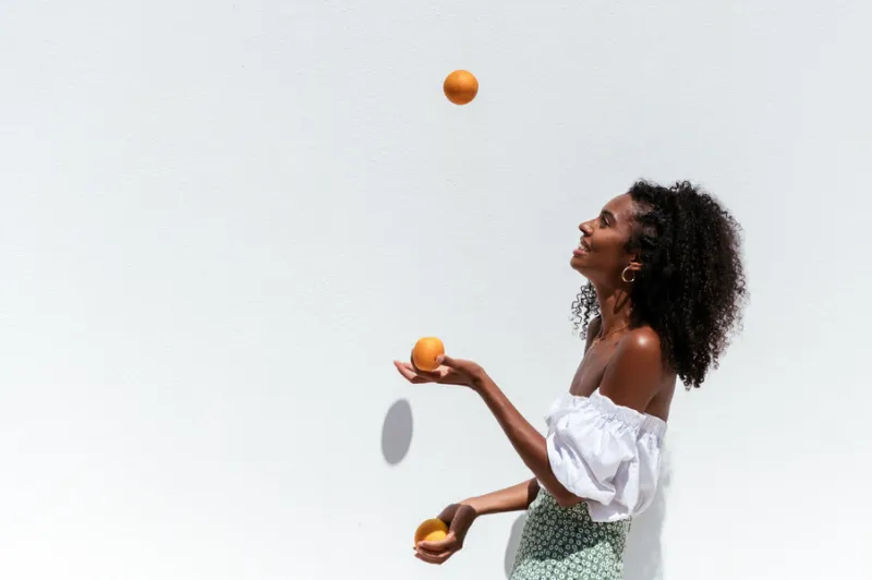 side view of happy young african american female in summer outfit juggling with ripe oranges against white wall in sunny day