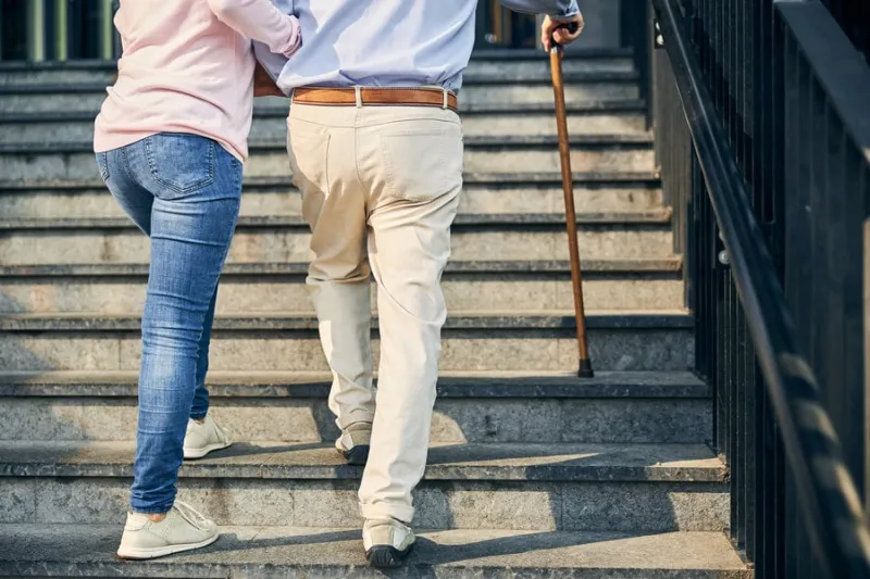 cropped back photo of a male with walking stick and female volunteer lifting up the stairs outdoors