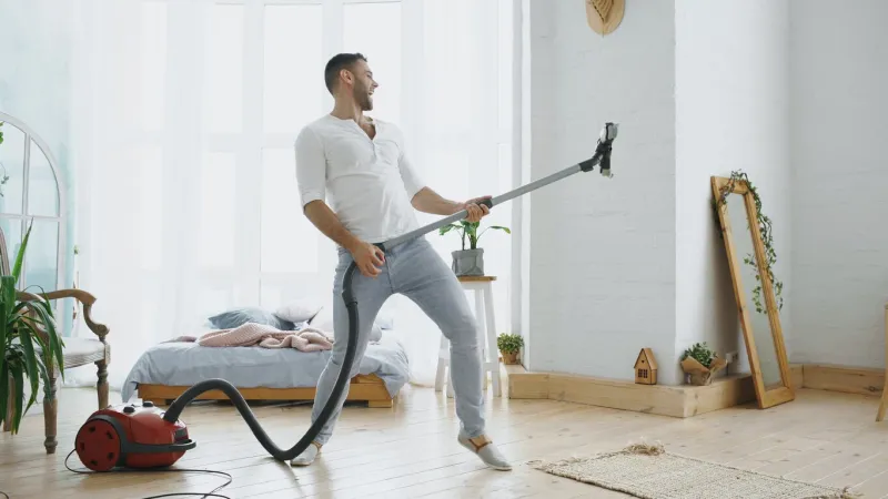 young man having fun cleaning house with vacuum cleaner dancing like guitarist