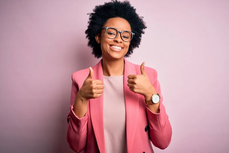 young beautiful african american afro businesswoman with curly hair wearing pink jacket success sign doing positive gesture with hand, thumbs up smiling and happy cheerful expression and winner gesture