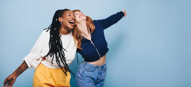 two happy best friends laughing and having a good time while embracing each other in a studio two cheerful female friends making fun memories while standing against a blue background