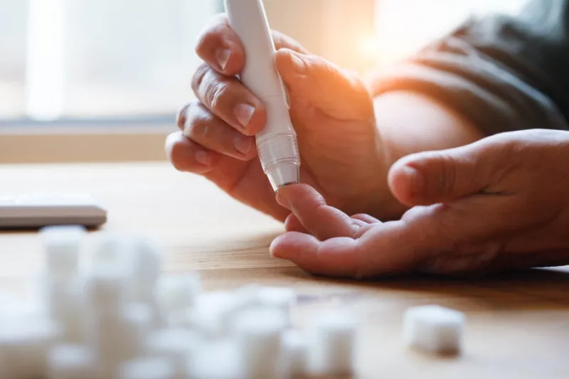 senior woman hands using lancet on finger at home to check blood sugar level, glucometer and sugar cubes on wooden table close up, diabetes concept, elderly health care, sunny morning