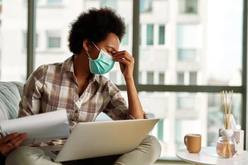 african american woman with face mask having a headache while going through paperwork and using laptop while working at home
