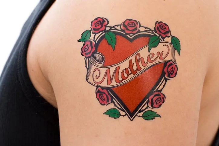 decorative tattoo on a male arm featuring the word mother on a scroll overlaying a red heart surrounded by pink roses with green leaves