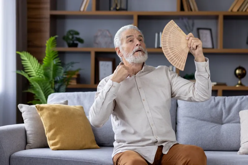 elderly caucasian man sitting on a gray sofa, using a handheld fan to cool off during a warm day indoors, looking uncomfortable and sweaty in a well-decorated living room