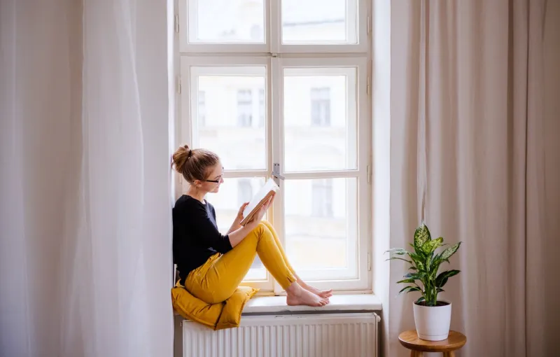 a young happy college female student with a book sitting on window sill at home, studying