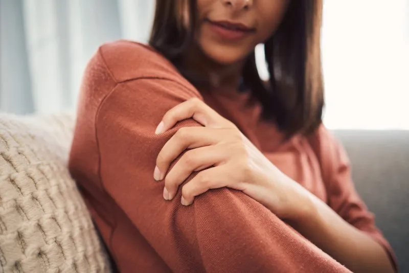 closeup shot of a mixed race woman in casual wear holding her arm while suffering pain and sitting on her sofa in the lounge at home one unrecognizable female struggling with an injury and arthritis