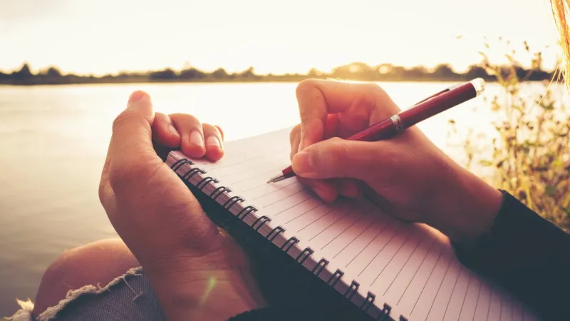 close up hand of young woman with pen writing on notebook at riverside in the evening