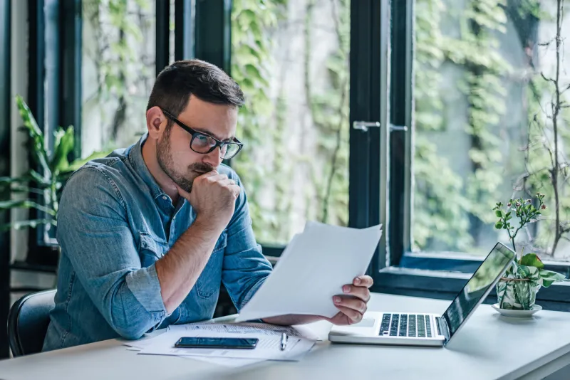 serious entrepreneur analyzing documents male professional reading reports while working on laptop he is sitting at table in home office