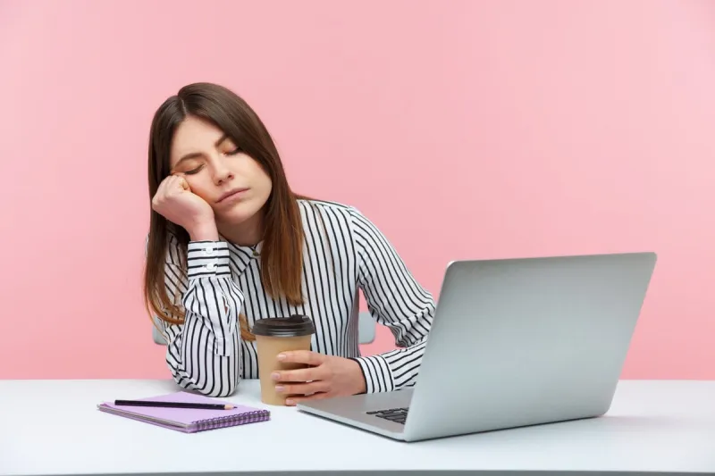 sleepy inefficient woman office worker napping leaning head on hand and holding coffee cup sitting at workplace with laptop, physical exhaustion indoor studio shot isolated on pink background