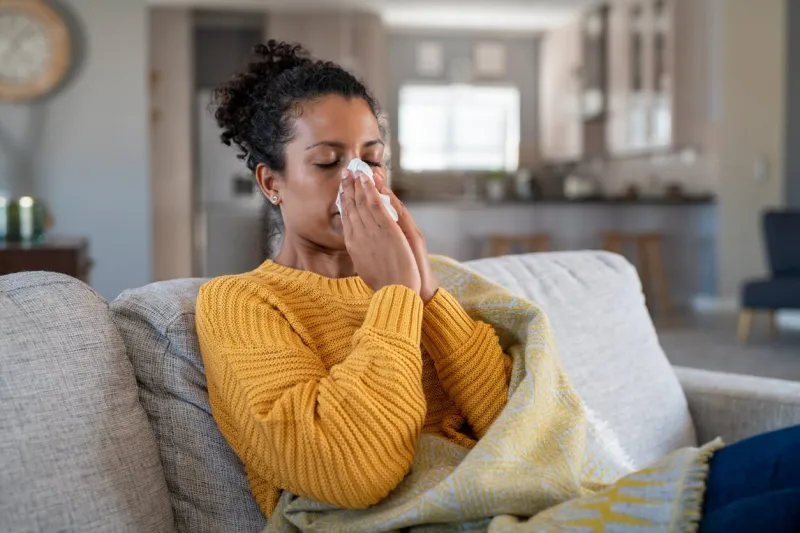 portrait of young black woman sneezing in to tissue at home sick african woman wrapped in blanket sitting on sofa blowing her nose at home ill girl sneezing with runny nose in winter
