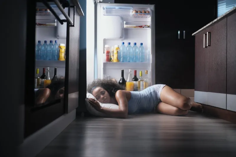 young hispanic woman suffering for summer heat and lack of air conditioning at home black girl covered with sweat sleeping on floor with head inside fridge