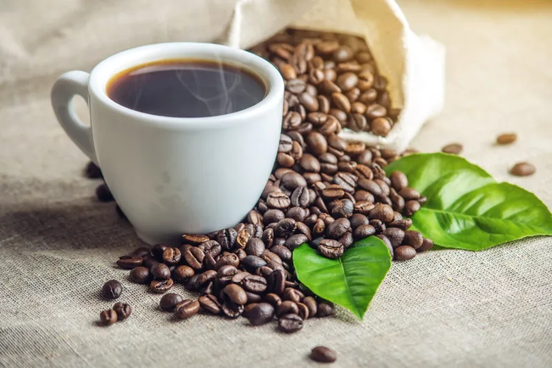 white black espresso cup with a pile of coffee beans and green leaves in bag on white linen background the concept of the production of fresh aromatic drink
