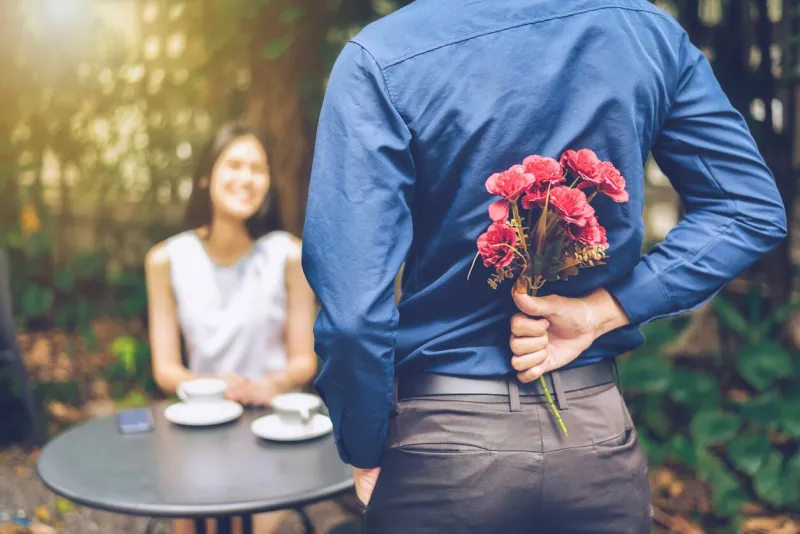 the man is hiding red flowers behind him in order to surprise his girlfriend