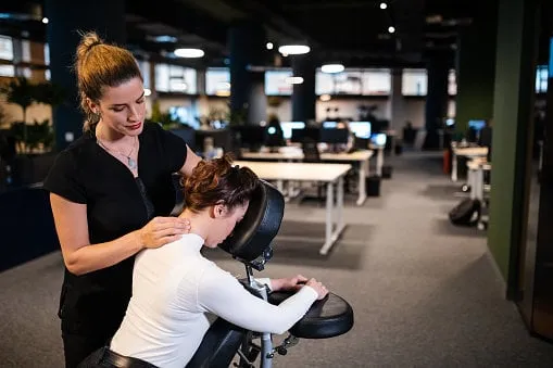 female massage therapist giving businesswoman massage for her back and neck pain, while she sitting in a mobile massage chair in her work office