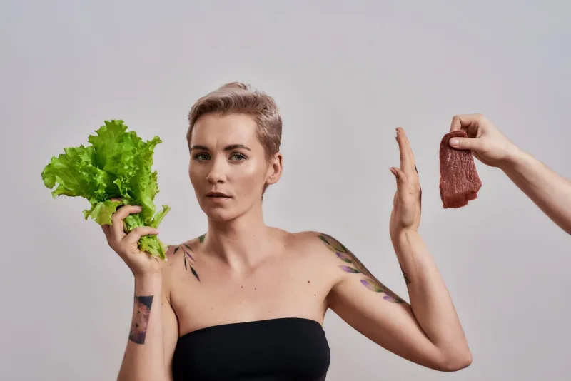 attractive tattooed woman with pierced nose refuses to eat meat offered to her, choosing fresh green lettuce isolated over grey background female with stop hand sign vegetarian nutrition concept
