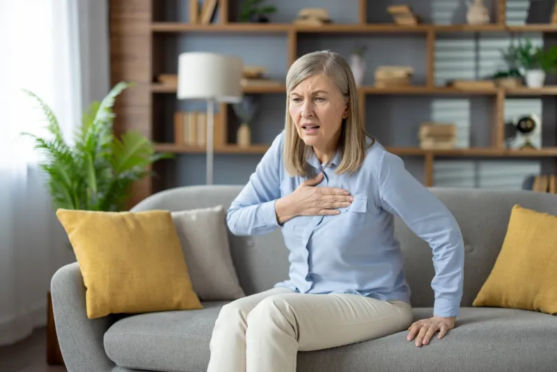 elderly female grabbing chest with palm while sitting on pillowed couch with shocked expression ill woman feeling sharp cardiac pain and risk of infarction while sitting alone in modern flat