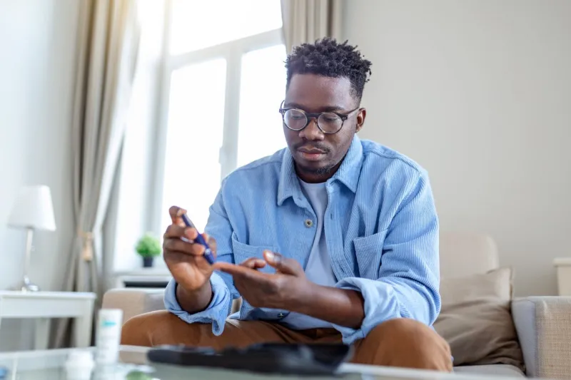 african man is sitting at the sofa and taking blood from his finger due to diabetes the daily life of a man of african-american ethnicity person with a chronic illness who is using glucose tester