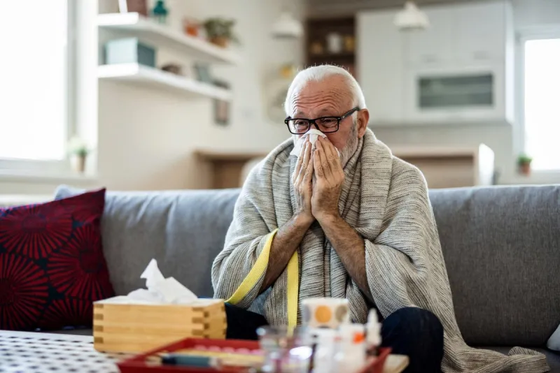 cold exhausted senior man with flu wrapped in a warm blanket blowing his nose with a tissue in the livingroom man with a cold lying in sofa holding tissues