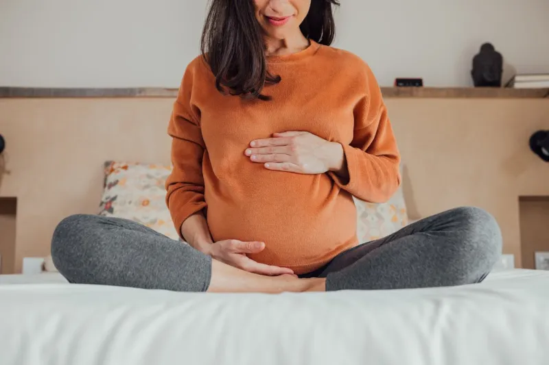 front view of cropped long hair brunet caucasian pregnant woman, sitting in crossed legs, in yoga pose, in bed at home, holding her belly with hand maternity, pregnancy, new life
