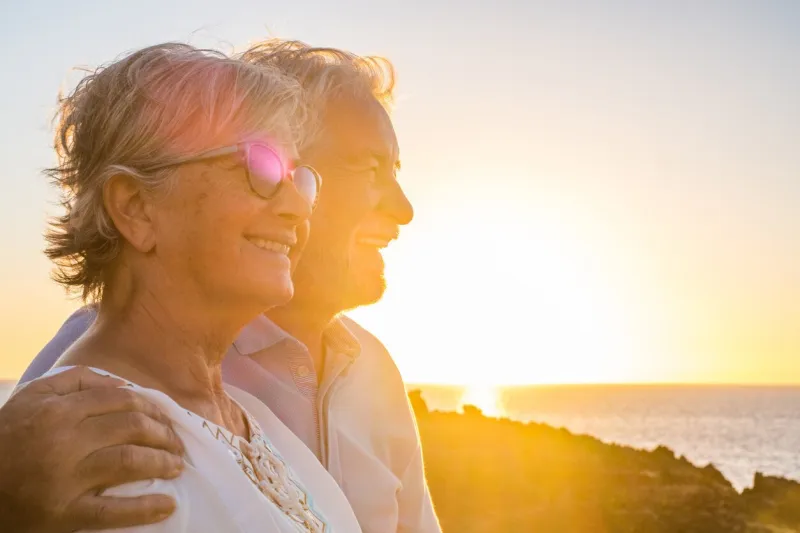 couple of two cute seniors together enjoying summer and having fun at the beach looking at the sea or ocean with sunset - mature people having a good lifestyle