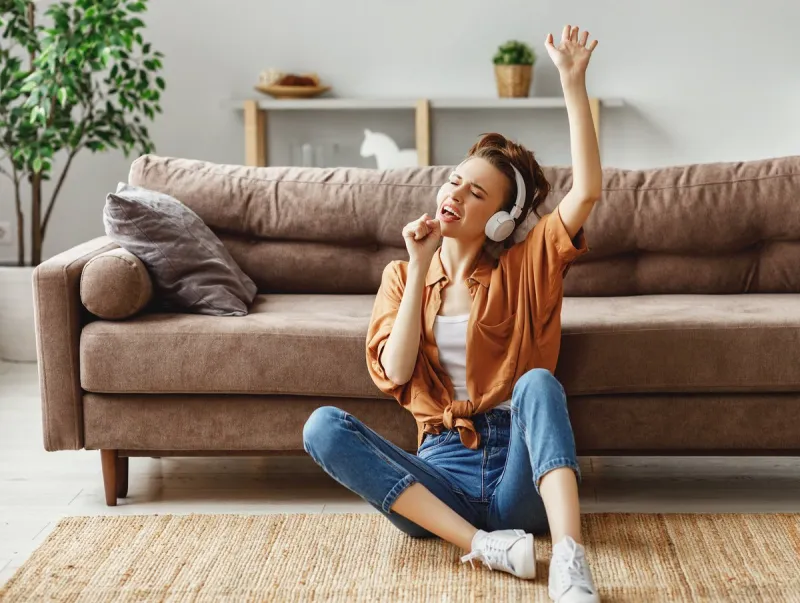 young female in casual outfit listening to music in headphones and singing while sitting on floor near sofa and having fun at home