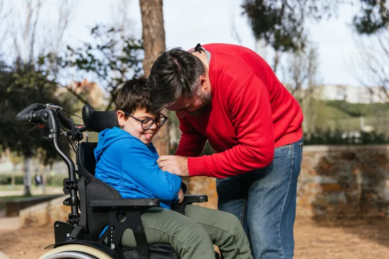 father playing with hisson with disability son in a wheelchair while they enjoy time together outdoors in the park parenthood, childhood and disabled people concept