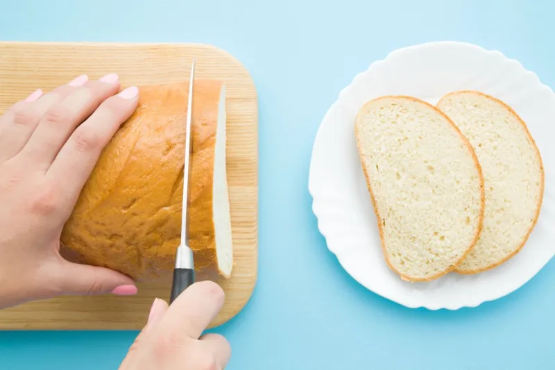 woman's hands cutting loaf of white bread with knife slices on plate on pastel blue desk preparing breakfast point of view shot closeup top view