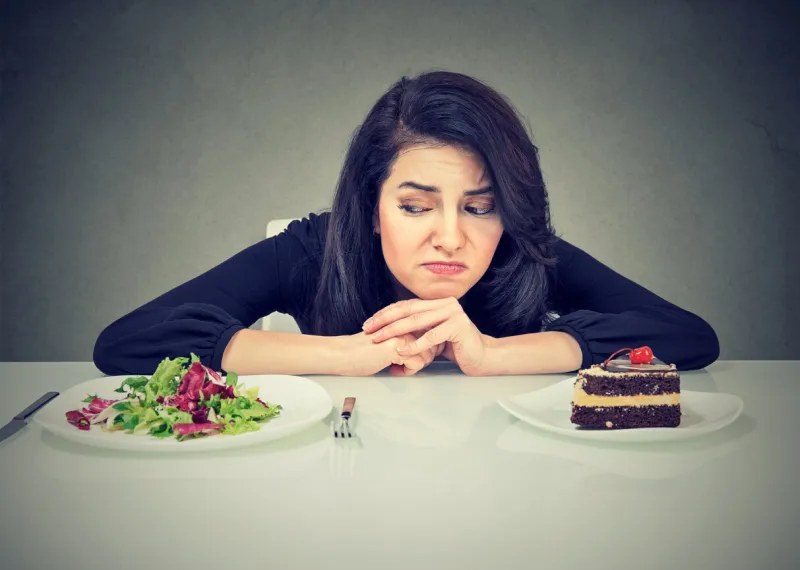 beautiful brunette sitting at desk and suffering from choice between healthy salad and sweet dessert